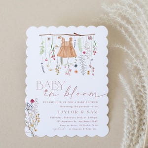 May include: A white baby shower invitation with a scalloped edge. The invitation features a watercolour illustration of a baby dress hanging from a branch with flowers. The text reads "Baby in Bloom" and includes details for the shower, including the date, time, and location.