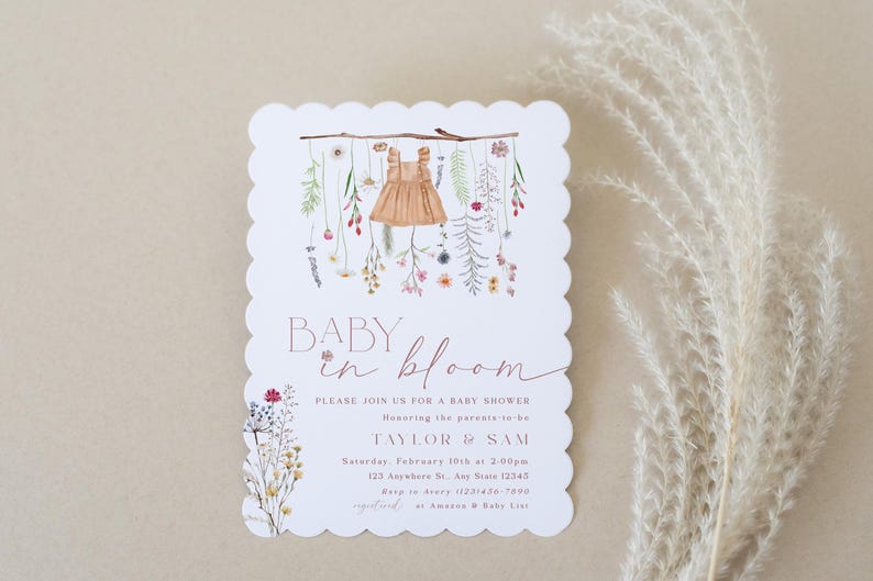 May include: A white baby shower invitation with a scalloped edge. The invitation features a watercolour illustration of a baby dress hanging from a branch with flowers. The text reads "Baby in Bloom" and includes details for the shower, including the date, time, and location.
