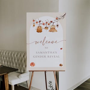 May include: A sign welcoming guests to Samantha's gender reveal party. The sign, displayed on an easel, features a watercolour design of baby clothes and autumn leaves. The words "welcome to Samantha's gender reveal" are written on the sign.