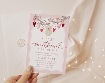 May include: Pink baby shower invitation with a watercolour illustration of a baby onesie hanging from a branch with pink and red hearts. The text reads "A little sweetheart is on the way".