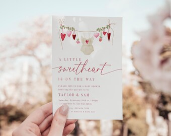 May include: A baby shower invitation with a watercolor illustration of a garland of pink and red hearts with a baby onesie hanging in the middle. The text reads "A little sweetheart is on the way" and includes the names "Taylor & Sam" and the date and time of the shower.