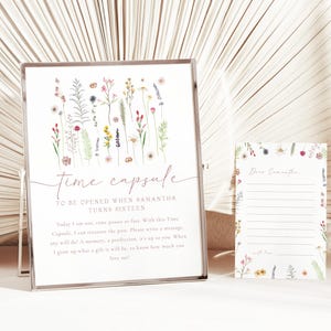 以下が含まれることがあります： 額縁に入ったタイムカプセルのサインと、それに合わせたカード。サインには「time capsule」と書かれており、その下にテキストがあり、水彩画の草花のイラストで飾られています。カードも草花のイラストで飾られています。