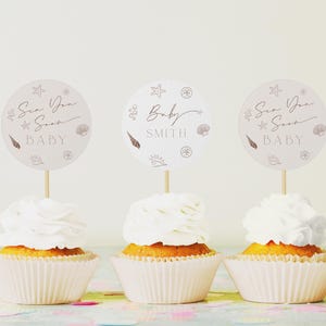 May include: Three cupcakes with white frosting and round toppers. The toppers have a seashell theme with the words "Sea You Soon BABY" and "Baby SMITH". The cupcakes are in white paper cups, ideal for a baby shower.