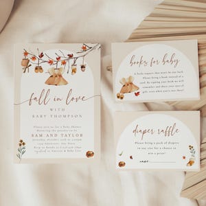 May include: A baby shower invitation set featuring a main card, a books for baby card, and a diaper raffle card. The cards are cream-colored with watercolor illustrations of autumn leaves, baby clothes, and text in brown script and serif fonts.
