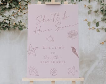 May include: A light pink baby shower welcome sign on a wooden easel. The sign reads "She'll be Here Soon" and "Welcome to Samantha's Baby Shower" with seashell and starfish illustrations. The background is a wall covered in green vines.