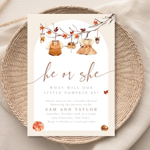 Peut inclure: Une invitation de révélation du sexe avec le texte "he or she" et "What will our little pumpkin be ?" Le design présente des illustrations à l'aquarelle de vêtements de bébé, de feuilles et de citrouilles. L'invitation est posée sur un panier tressé.
