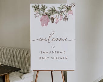 May include: A sign welcoming guests to Samantha's baby shower. The sign, on a wooden easel, displays a watercolour illustration of baby clothes and the word "welcome". The sign is decorated with greenery and flowers.