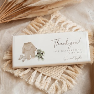 May include: A white chocolate bar with a thank you message for celebrating with Sam and Taylor. The bar features a watercolor illustration of a baby in a brown onesie, socks, and a pine branch.