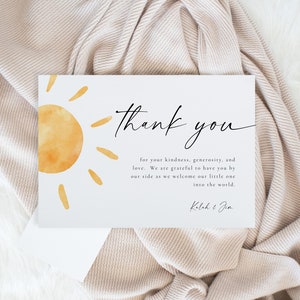 May include: A white thank you card with a watercolor illustration of a yellow sun. The text reads "thank you for your kindness, generosity, and love. We are grateful to have you by our side as we welcome our little one into the world. Kalah & Jim."