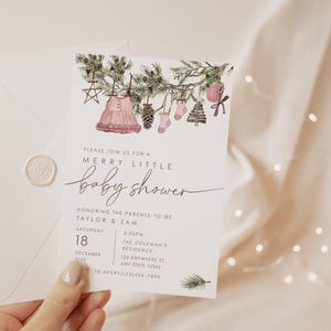 Editable holiday baby shower invite featuring pine garland with hanging baby clothes, star ornament, pinecone, and pink accents. Modern Christmas baby shower template.
