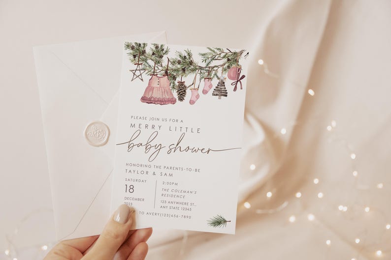 Editable holiday baby shower invite featuring pine garland with hanging baby clothes, star ornament, pinecone, and pink accents. Modern Christmas baby shower template.