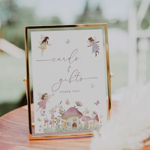 May include: A framed sign with a gold border, displaying the words "cards & gifts" in elegant script, along with illustrations of fairies, mushrooms, and butterflies. The sign reads "Thank You!" at the bottom. The sign is set on a wooden surface.