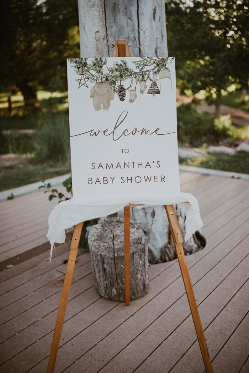 May include: A white sign with a brown wooden easel, decorated with a watercolour illustration of a pine branch with ornaments, reads "Welcome to Samantha's Baby Shower".