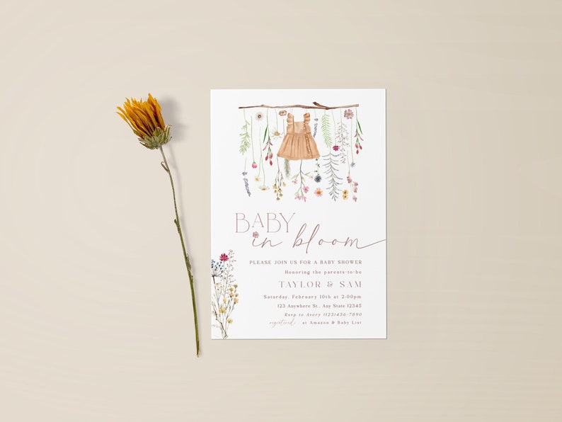 May include: A baby shower invitation with a floral theme. The invitation features a watercolour illustration of a baby dress hanging from a branch with flowers and greenery. The text reads "Baby in Bloom" and includes the names "Taylor & Sam" and the date and time of the shower.
