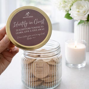 May include: A glass jar containing wooden tokens, with a lid inscribed with "Identity in Christ." A lit candle and a vase of white flowers are nearby. The tokens are for learning, living, and sharing scripture. The jar is in cm.