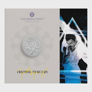 Puede incluir: Una moneda de plata con Freddie Mercury y el texto "FREDDIE MERCURY" en amarillo. La moneda está sobre un fondo gris claro con el logotipo de la Royal Mint. Un objeto de colección.