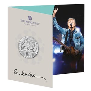 May include: A silver coin featuring a musical note design with the signature "Paul McCartney" and the text "The Royal Mint" on a white card with a photo of Paul McCartney performing on stage.