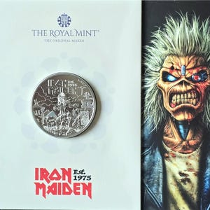 Puede incluir: Una moneda de plata con la inscripción "Iron Maiden Est. 1975", presentada en una tarjeta blanca con el logotipo "The Royal Mint". La moneda presenta una ilustración detallada. Un gráfico de un monstruo está a la derecha. Objeto de colección.