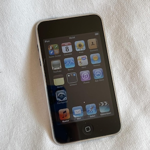 Könnte beinhalten: Ein schwarzer iPod touch mit silbernem Rahmen, der verschiedene App-Symbole auf seinem Bildschirm anzeigt. Der Bildschirm zeigt die Uhrzeit 10:44 Uhr an. Das Gerät liegt auf einer weißen, strukturierten Oberfläche.