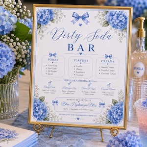 May include: A rectangular Dirty Soda Bar sign in a gold frame featuring blue hydrangea illustrations, a bow graphic, and text listing sodas, flavors, creams, and combinations. The sign is displayed on a gold easel with blue floral accents and bottles.