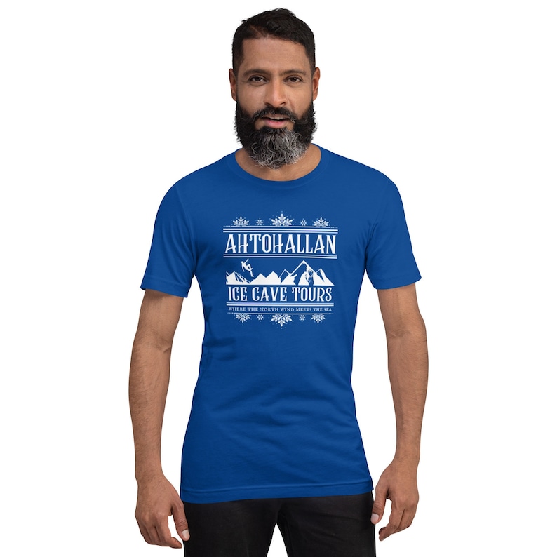 Puede incluir: Una camiseta azul real con un dise&ntilde;o gr&aacute;fico blanco que dice "Ahtohallan Ice Cave Tours" con un borde de copos de nieve y una escena de monta&ntilde;a con un esquiador.