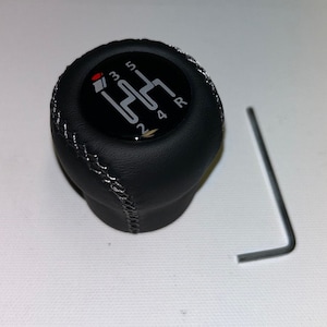 May include: Black leather gear shift knob with a silver stitching detail. The knob has a black and silver circular design with the numbers 1, 2, 3, 4, 5, and R printed on it.