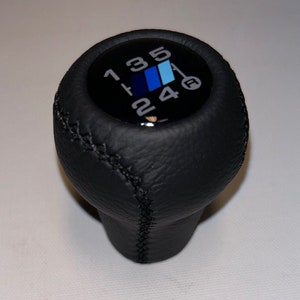Pode incluir: Manípulo da alavanca de câmbio em couro preto com números brancos e um emblema azul, azul claro e azul escuro. O padrão de mudança é 1, 3, 5, R, 2 e 4. O manípulo tem uma costura.