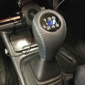 May include: A black leather gear shift knob with blue stitching and a black and blue gear shift pattern with the numbers 1, 3, 5, R, 2, 4, and 6.