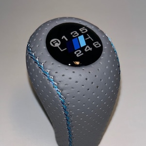 May include: Grey leather gear shift knob with blue stitching and a black circular gear shift pattern with the numbers 1, 3, 5, R, 2, 4, and 6.