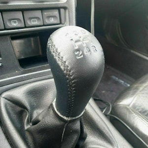 Op de afbeelding: Een zwarte leren versnellingspookknop met een gestikt patroon en de nummers 1, 2, 3, 4, 5 en R erop gedrukt. De knop is bevestigd aan een zwarte leren versnellingspookhoes.