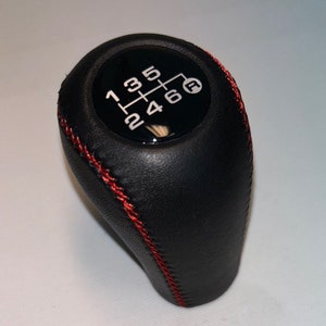 Peut inclure: Pommeau de levier de vitesses en cuir noir avec surpiqûres rouges et un motif de vitesses noir et argent avec les chiffres 1, 2, 3, 4, 5, 6 et la lettre "R" pour la marche arrière.