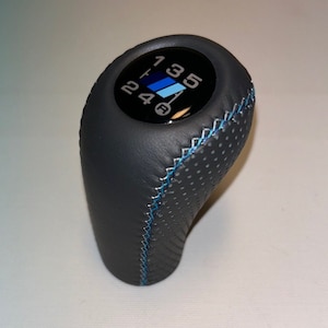 Op de afbeelding: Een zwarte leren versnellingspookknop met blauwe stiksels en een zwart en blauw versnellingschema. De knop heeft een gestructureerd oppervlak.