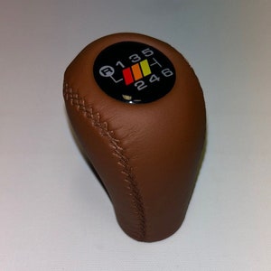 Peut inclure: Pommeau de levier de vitesses en cuir marron avec un dessus circulaire noir présentant un motif de changement de vitesse blanc avec des rayures rouges, orange et jaunes. Les chiffres 1, 3, 5, R, 2, 4 et 6 sont imprimés en blanc sur le cercle noir.