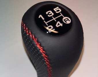 Può includere: Pomello del cambio in pelle nera con cuciture rosse e una parte superiore in plastica nera con numeri bianchi 1, 2, 3, 4, 5 e R.
