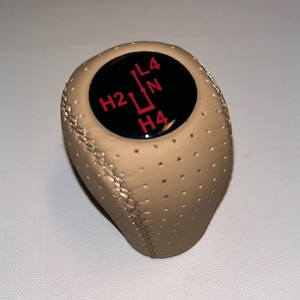 May include: Tan leather-wrapped gear stick knob with a black top displaying red lettering and a gear shift pattern. The knob has a perforated design and visible stitching along the side.