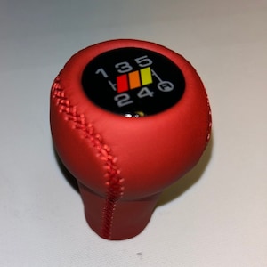 Peut inclure: Pommeau de levier de vitesse en cuir rouge avec un dessus circulaire noir présentant un schéma de vitesses à 5 rapports avec des rayures rouges, orange et jaunes.