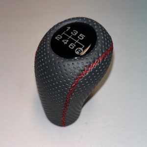 Może przedstawiać: Czarny skórzany gałka zmiany biegów z czerwonymi szwami i czarnym wzorem biegów na górze. Liczby 1, 2, 3, 4, 5 i 6 są widoczne na wzorze biegów.