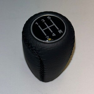 Könnte beinhalten: Schwarzer Leder-Schalthebelknauf mit einem silbernen 5-Gang-Schaltmuster und dem Buchstaben 'R' für den Rückwärtsgang.