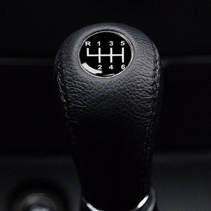 Peut inclure: Un pommeau de levier de vitesses en cuir noir avec une bague chromée et un motif de vitesses blanc avec les chiffres de 1 à 6 et R.