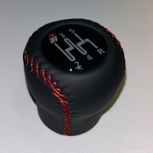 May include: Black gear shift knob with red stitching. The top features a diagram of the gear pattern in white, with numbers and an "R" for reverse. The knob is made of a smooth, possibly leather-like material.