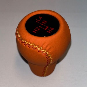 Może przedstawiać: Pomocnik zmiany biegów z pomarańczowej skóry z czarnym wierzchołkiem i czerwonym napisem "H2 L4 N H4".