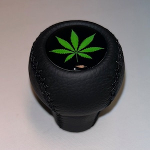 Op de afbeelding: Zwarte leren versnellingspookknop met een groen marihuanabladontwerp op de bovenkant.
