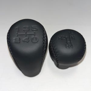 Könnte beinhalten: Zwei schwarze Lederschaltknäufe. Der größere Knauf hat die Zahlen 1, 2, 3, 4, 5 und R eingeprägt. Der kleinere Knauf hat H2, N und H4 eingeprägt. Beide Knäufe sind genäht.