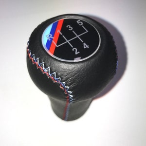 Puede incluir: Pomo de palanca de cambios de cuero negro con un logotipo BMW blanco, azul y rojo y un patrón de cambio de 5 velocidades.