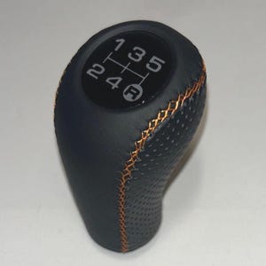 May include: Black leather gear shift knob with orange stitching. The top features a diagram of the gear positions: 1, 3, 5, 2, 4, and R (reverse). The knob has a textured surface for grip.