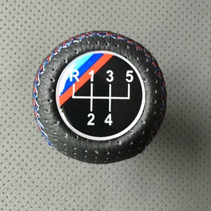 Peut inclure: Pommeau de levier de vitesses en cuir noir avec une surface perforée et un logo BMW blanc, bleu et rouge avec les chiffres 1, 2, 3, 4, 5 et R.