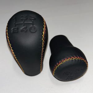 Pode incluir: Dois manípulos de mudança de velocidades pretos com acabamento mate liso e um padrão de mudanças em relevo no topo. Cada manípulo é decorado com costuras em cruz com fio vermelho e amarelo alternado ao longo das bordas curvas.