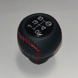 May include: Black gear shift knob with red stitching. The top of the knob has a diagram of the gear shift pattern, with numbers 1 through 5, and R for reverse. The knob is made of a textured material.