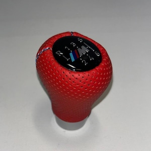 May include: A red, perforated leather shift knob with a black top. The top features the gear shift pattern and the BMW M logo in blue, black, and red. The knob is designed for a car's manual transmission.