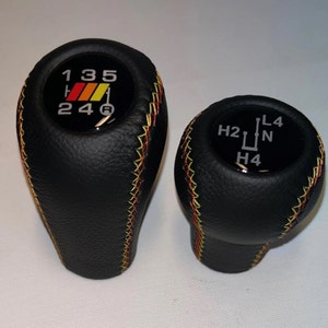 May include: Two black leather gear shift knobs with yellow stitching. The larger knob has a black and red sticker with the numbers 1, 3, 5, 2, 4, and a symbol. The smaller knob has a black sticker with the letters H2, L4, N, and H4.